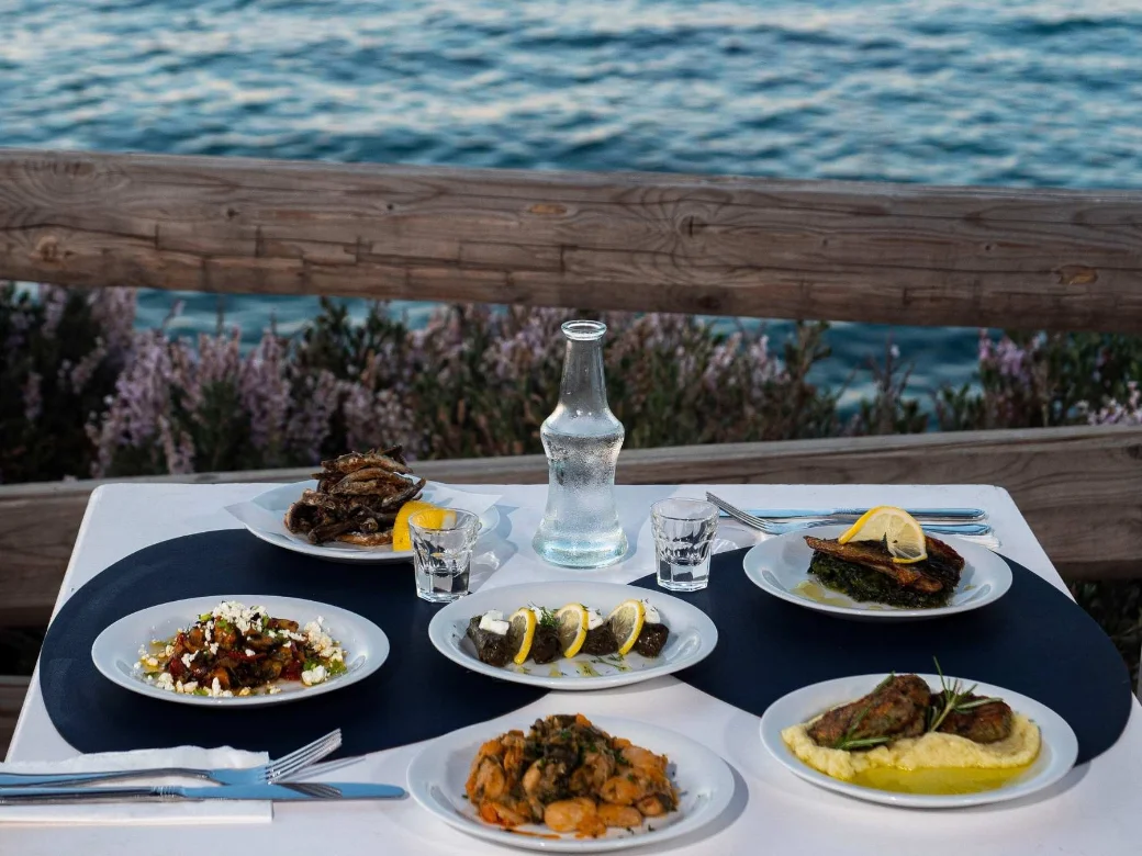 Six plates of the day laid out on a seafront table with a chilled carafe of raki