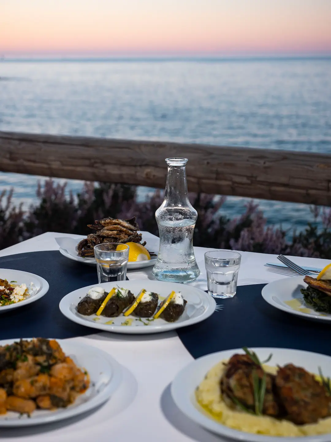 A full table of meze — dolmades, fried marides, shrimp and keftedes — with a frosted carafe of raki, the sea and a pink dusk sky beyond the terrace rail